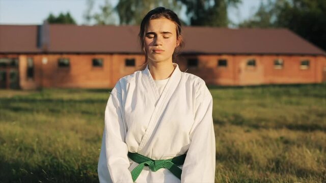 Young karate girl in a white kimono and a green belt meditates sitting on the grass in nature. Front view. Close-up. Slow motion. Blurred background