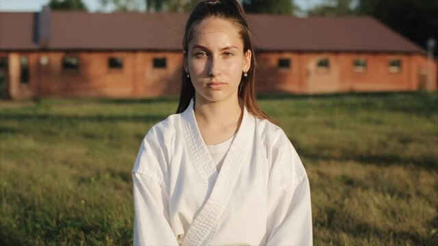 A young woman in karate training meditates sitting on the grass with her eyes closed and then opening them. Front view. Close-up. Slow motion. Camera zooms in
