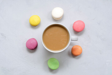 A cup of coffee with milk in the center and multi-colored macarons around it on a light concrete background with a place for text. Flat lay, top view.