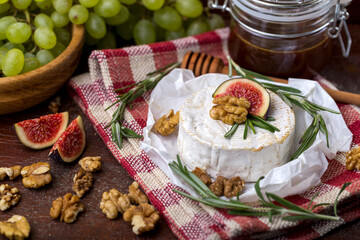 Camembert cheese on red and white checkered towel, on paper, pelled walnuts, sprigs of rosemary, green grapes, honey in jar,figs slices. Brown wooden background.Gorizontal format.