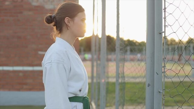 A girl athlete in a white kimono stands in profile against the sunset and then shouts showing the reception of karate. Close-up. Slow motion