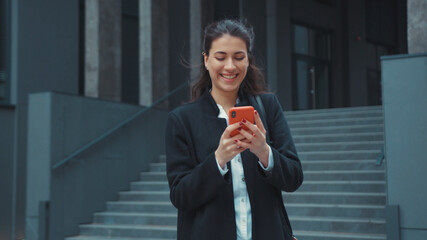 Young cheerful office girl caucasian pretty woman using smartphone networking social media while walking in business district outdoors. Carefree. Urban lifestyle.