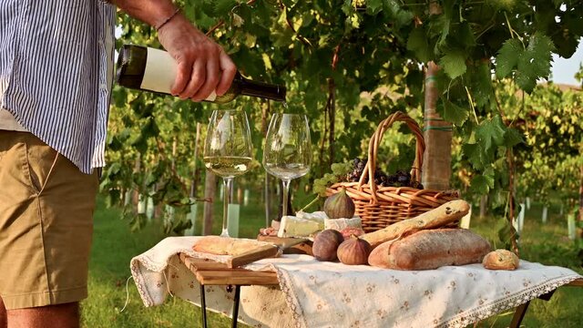 The man pours white wine into glasses. Beautiful people having romantic lunch with lots of tasty food and wine, sitting together on the picnic blanket at the vineyard on a sunny day. 4k footage