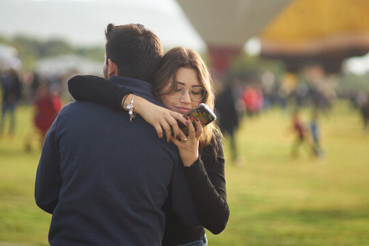 Young couple hugging at a festival. The woman is distracted on her phone
