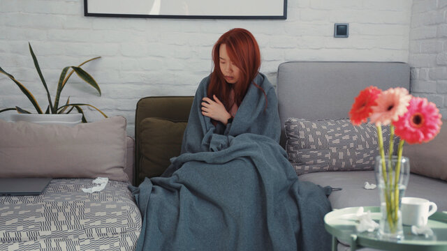 Unhealthy cold asian woman shivering wrapped up in blanket trying to warm up on sofa in the living room. Staying home. Symptoms of flu. Pandemic. Coronavirus.