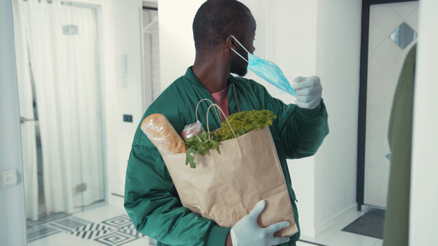 African American Man Entering Home With Food Package, Taking Off Surgical Mask. Handsome Guy Coming Back Home From Work. Casual Lifestyle. Routine. Health Care. Quarantine.