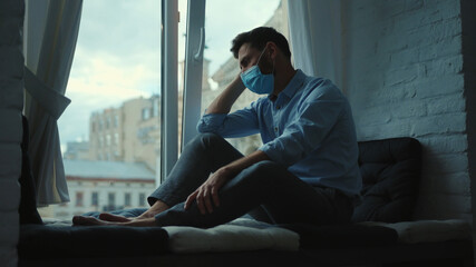 Upset businessman in medical mask at home sitting on windowsill and looking at street on quarantine COVID-19 pandemic epidemic depressed portrait slow motion