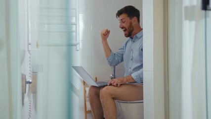 Smiling man workaholic is working in toilet on computer happy reading good news on laptop at home businessman positive success communication table modern slow motion