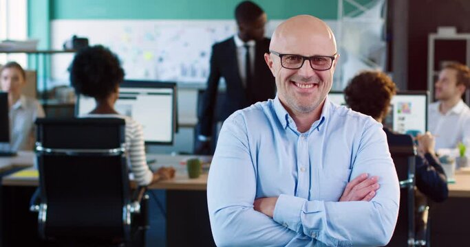 Portrait of handsome bald business worker with glasses sitting in office with arms crossed on his chest and laughing.