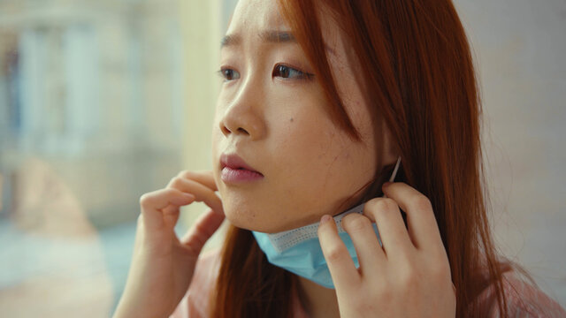 At sunlight asian woman remove medical mask at home sitting on windowsill and looking at street on quarantine COVID-19 pandemic epidemic depressed portrait slow motion