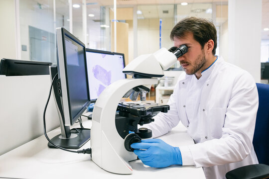 Man in white apparel and gloves examining samples with microscope while working in innovative laboratory.