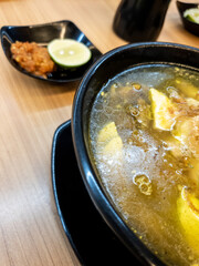 soup served in a black bowl complete with vegetables, seasonings, spoons & forks, asian style food served on a wooden table.
