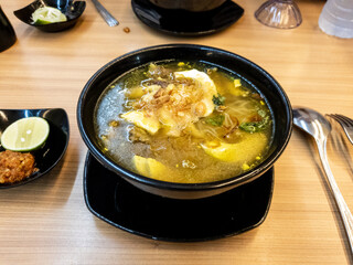 soup served in a black bowl complete with vegetables, seasonings, spoons & forks, asian style food served on a wooden table.