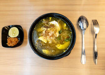 soup served in a black bowl complete with vegetables, seasonings, spoons & forks, asian style food served on a wooden table.