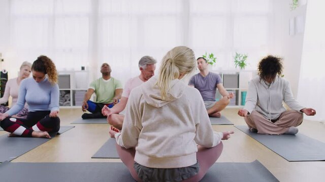 View from behind of group with teacher sitting on exercise mats stretching in yoga class inside community center - shot in slow motion