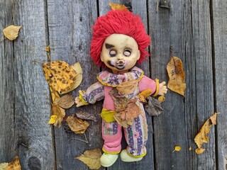 scary and old battered clown doll with burnt face and red hair on the background of old wooden board with autumn leaves