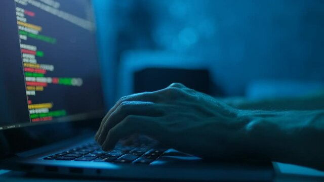 Hacker is working on laptop at night. Hands are typing a virus code at keyboard or hacking website. Closeup