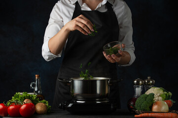 Close-up view of the professional chef in black apron adding chopped parsley in boiling water for soup on dark blue background. Backstage of preparing healthy meal. Food concept. Frozen motion.