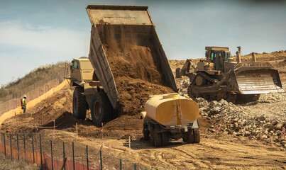 A very large haul dump truck at a construction site dumping a massive amount of dirt near a water...