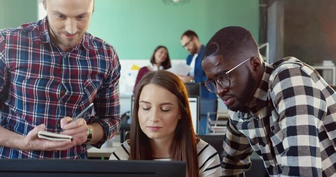 Three young people: two men and one woman, working at computer in coworking. Multinational team discusses project in office.