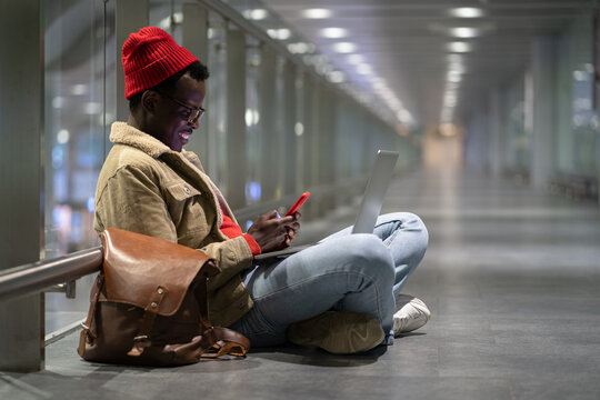 Side View Of Stylish African American Traveler Millennial Man Resting, Sitting On The Floor In Empty Hall Of Airport At Night, Using Phone, Chatting In Social Media, Waiting For A Flight And Boarding