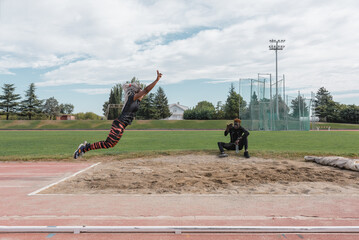 Ethnic female athlete making energetic leap into sandpit near trainer