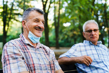 Fototapeta premium Two senior caucasian men sitting on bench in park in autumn or summer day - selective focus on man with mustaches smiling - real people leisure activity social distance concept side view copy space