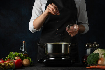 Close-up view of the professional chef in black apron salting water before cooking soup on dark blue background. Backstage of preparing meal. Healthy dish. Food concept. Frozen motion.