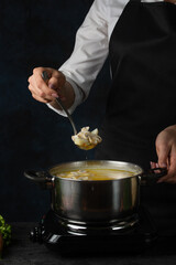 The chef in black apron cooking tasty soup in big pot on the stove. Dark blue background. Food concept.