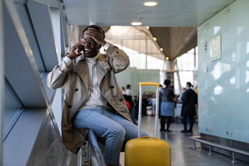Stressed Black man in spectacles shocked with bad news, talking on mobile phone, sitting in airport, makes facepalm by hand and forgot something important at home. Forgetfulness before the flight - obrazy, fototapety, plakaty