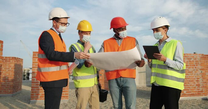 Group of mixed-races men and woman constructors in helmets and medical masks standing at building top and discussing constructing plan draft with tablet device. Coronavirus. Engineers and architectors