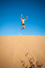 Youth jumping off a sand dune shirtless with hat and sunglasses on a clear day cloudless sky