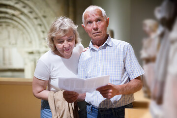 Male and female pensioners visiting exposition of museum and reading guidebook