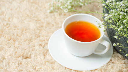White cup with tea placed on a cream carpet. Decorated with white flowers in the corner. 