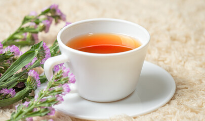 White cup with tea placed on a cream carpet. Decorated with purple flowers in the corner. 