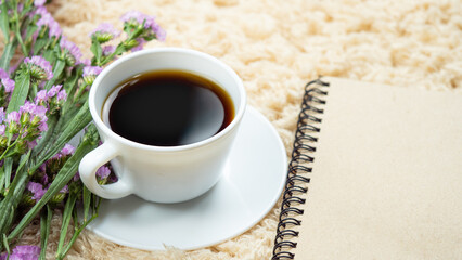 White cup with black coffee placed on a cream carpet. Decorated with white flowers in the corner of the coffee and pink notepaper.