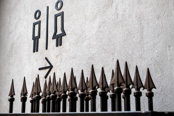 Black icon toilet sign with arrow on white wall and black wrought iron gate with pointed rusty spears below.
