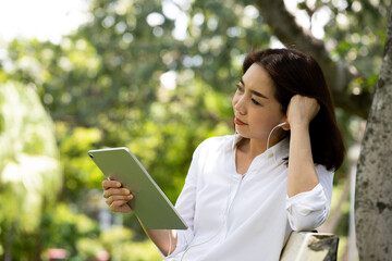 Portrait of a smiling young business woman using tablet for watching movies and listening music in the park with earphones. Changing workplace to create creative ideas and new ideas.