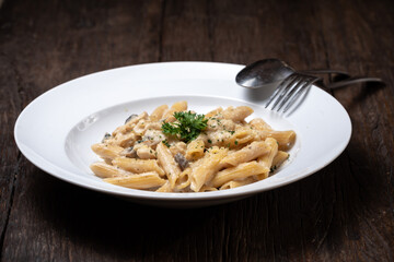 Homemade delicious pasta macaroni with white mushrooms chicken meet mushrooms in creamy cheese sauce in the white plate on the dark brown wood table. Parsley, spicy. Yellow, green.