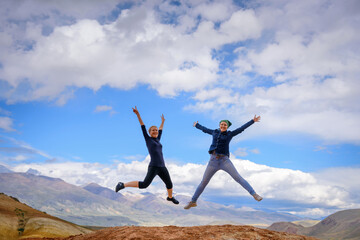 Fototapeta na wymiar Two young happy girls jump raising hands up with spectacular view of mountains. Female travellers full of joy against the mountains and blue sky with white clouds. Lifestyle in motion concept.