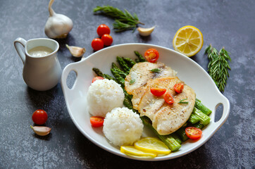 Fried tilapia, asparagus, cherry tomatoes, lemon and rice balls on a white plate.	
