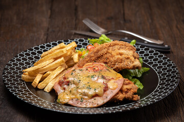Fried chicken with french fries and sausage, cheese, salad, greens - junk food and unhealthy food. Fast food set fried chicken and french fries. Yellow sauce. Black plate on the dark wooden table.