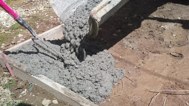 Truck pours wet mushy heavy concrete through chute in corners and side of campground campsite as man rakes, pushes, pulls and directs flow of cement in flat pad site ground, handheld close up pan