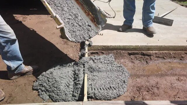 Large industrial cement truck pours wet concrete through feeder and discharge chute into formed border flat pad in RV campground site as blue collar men rake, level and spread concrete, close up pan