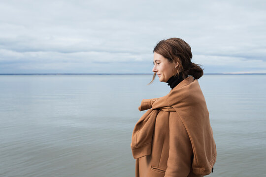 Portrait of a beautiful young model  in  a beige wool coat   smilling,  posing  against sea  in  sunny autumn day . Autumn warm photo. Woman smiling and look at camera, joyful  mood.