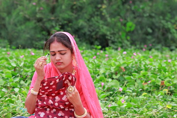 close-up portrait of An Indian young attractive stylish blonde woman sitting in the garden applying a color to her eyebrow. fashion model wearing style traditional pink rajputani shot dress, Make-up