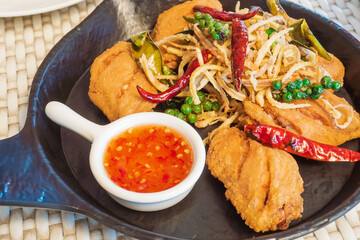 Fried chicken wings on a black plate, served with garnish with freshly ground pepper, dried chilli, and sweet spicy sauce. Appetizer Thai style.