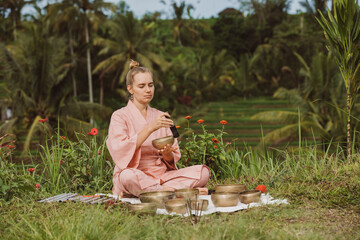 Blonde woman in a pink kimono playing on meditative Himalayan singing bowl. Sound meditation, sound healing. She is sitting on a green grass in Jatiluwih rice terraces. 