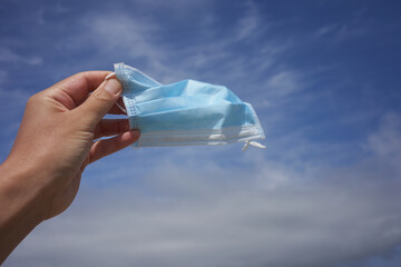 Hand holding a disposable surgical mask in the wind against the clouds and sky.