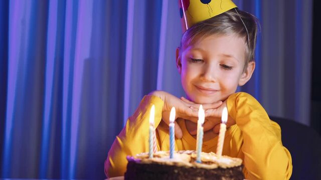 Happy child looks at a cake with candles and makes a wish.  Cute boy blows out candles on a cake on his birthday.  Child looks at the cake and makes a birthday wish. Boy celebrates his birthday.
